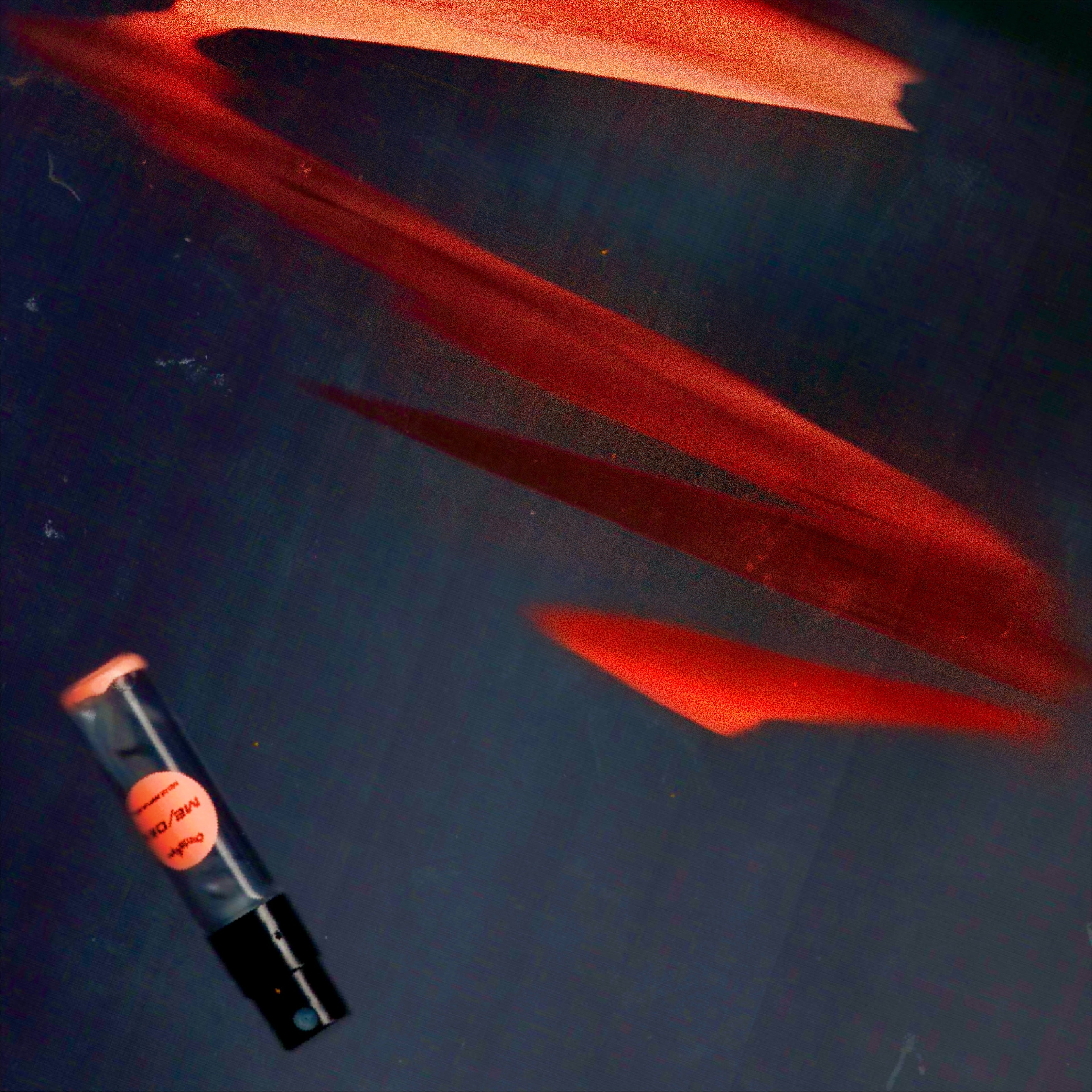 2mL spray sample of Chronotope Perfume’s Me/dea Eau de Parfum with Safety Orange label and matching enameled tip, positioned diagonally on a dark gray surface.