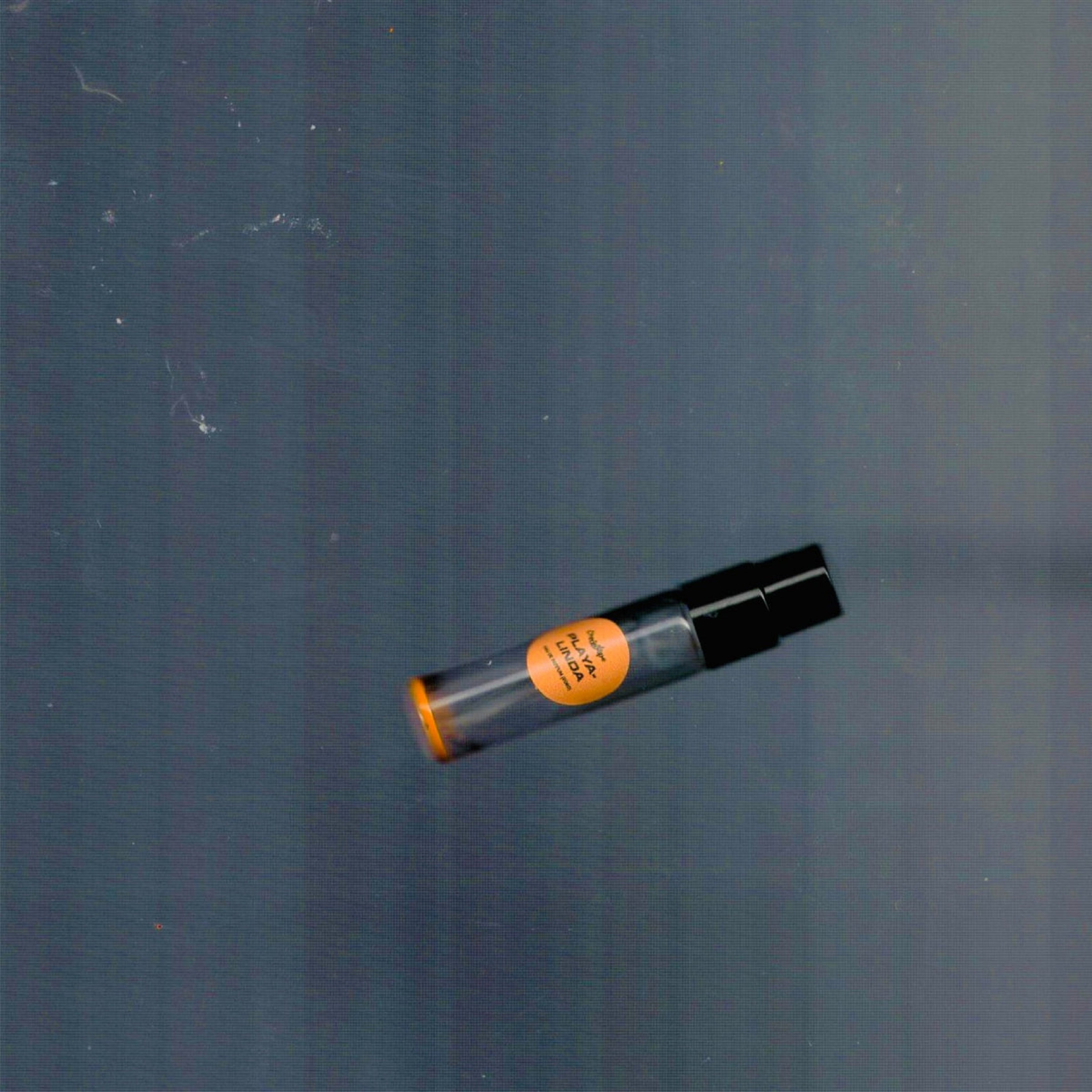 2mL spray sample of Chronotope Perfume’s Playalinda Eau de Parfum with orange label and matching enameled tip, positioned diagonally on a muted gray surface.