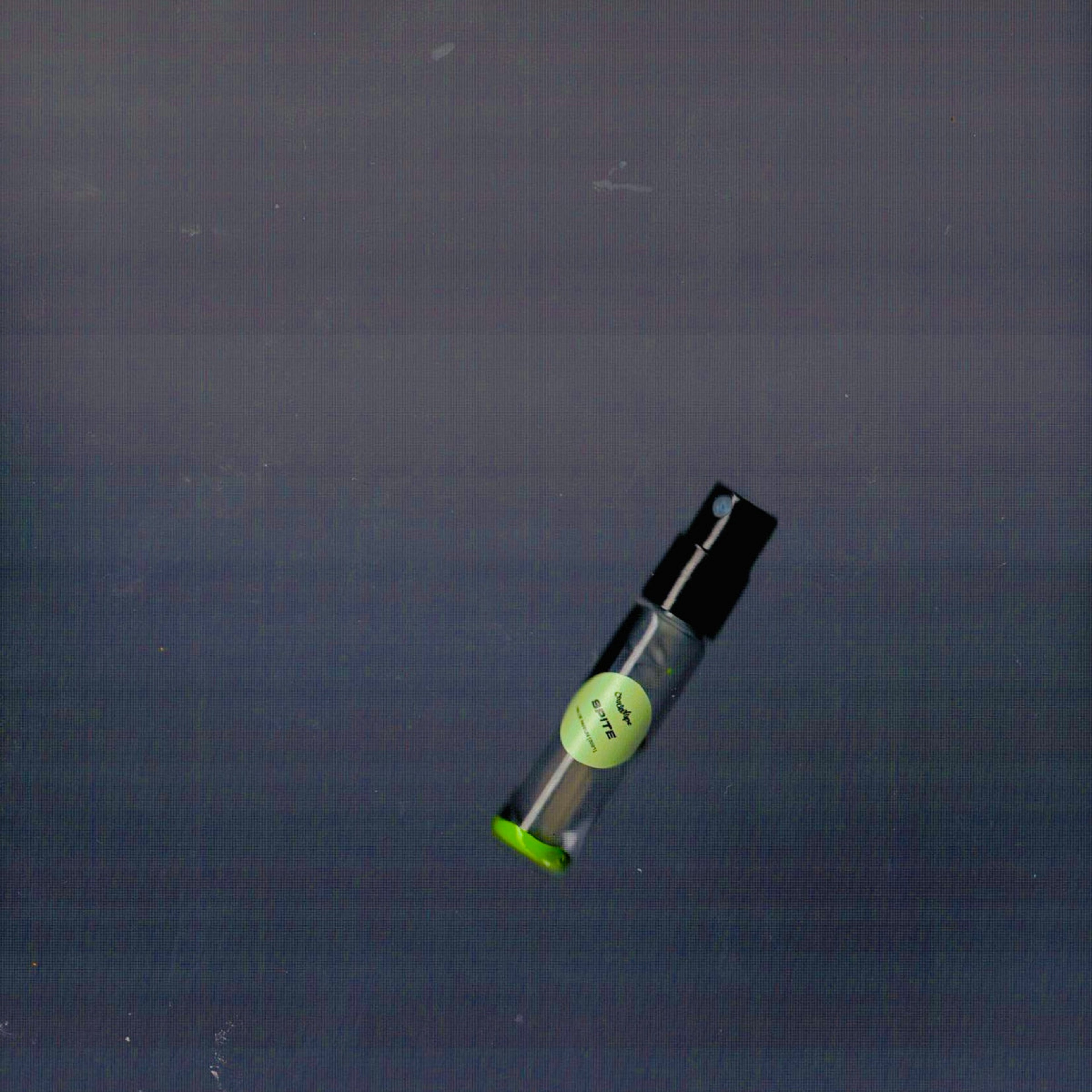 2mL spray sample of Chronotope Perfume’s Spite Eau de Parfum with green label and matching enameled tip, resting on a charcoal gray surface.