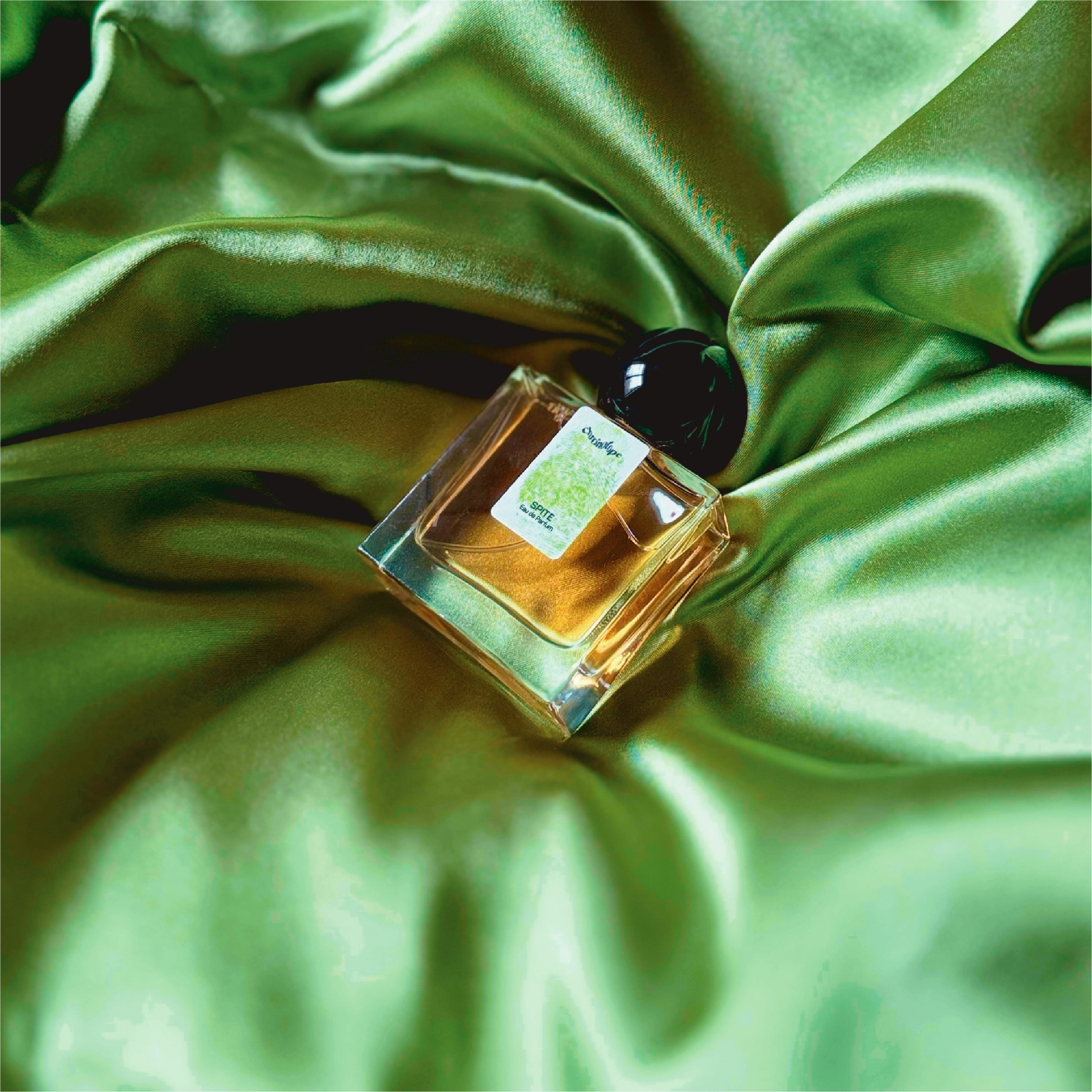 Nestled in a nest of green, silky, shining fabric with brilliant highlights and folds that appear black is a 30mL bottle of Chronotope Perfume’s Spite Eau de Parfum: A Perfume about Adaptation.