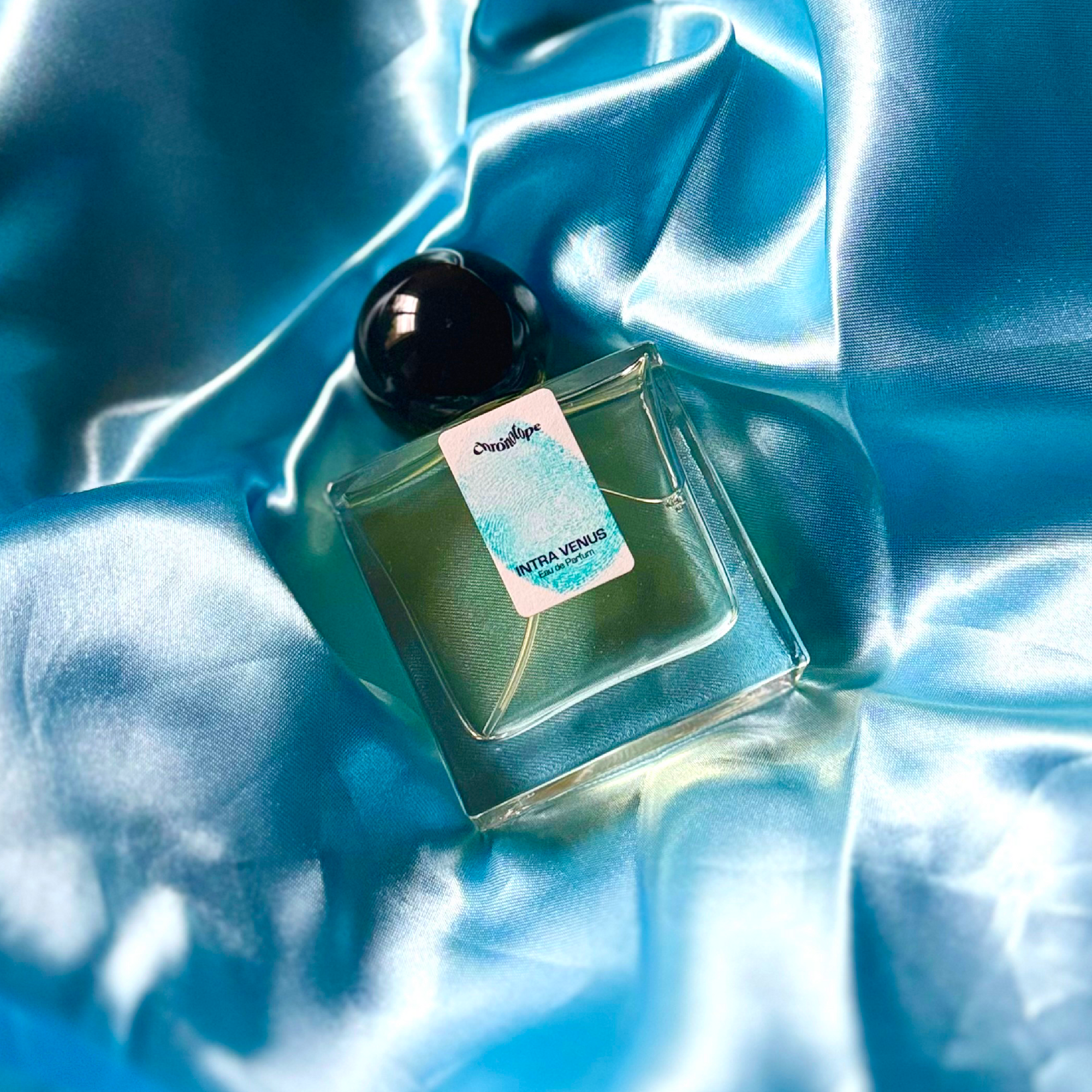A full, 30mL bottle of Intra Venus Eau de Parfum by Chronotopeis nestled into a nest of colored, silky, shining fabric that matches the color of the thumbprint on the bottle’s label.