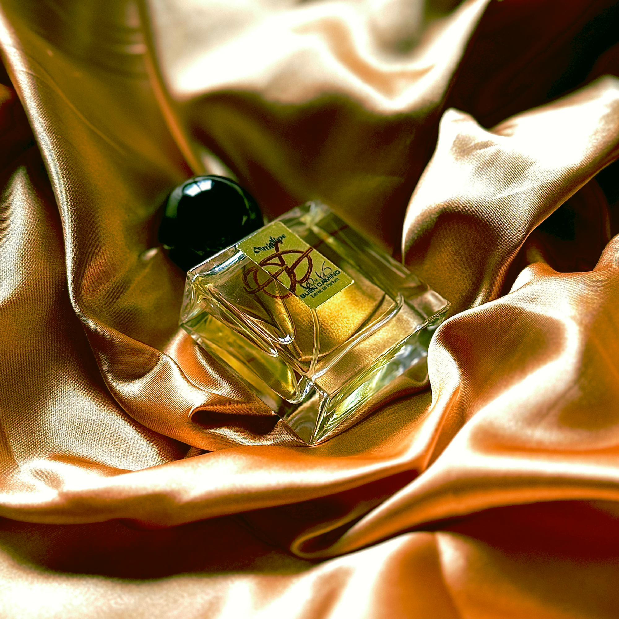 A bottle of Buen Camino Extrait de Parfum by Chronotope Perfume, nestled into shimmering gold fabric that reflects the color of the gold label on the face of the bottle.