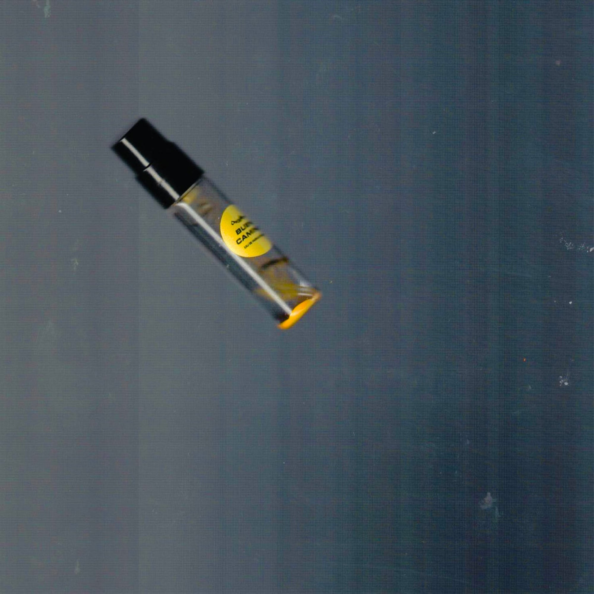2mL spray sample of Chronotope Perfume’s Buen Camino Eau de Parfum with Pilgrim Yellow label and matching enameled tip, resting on a dark gray background.