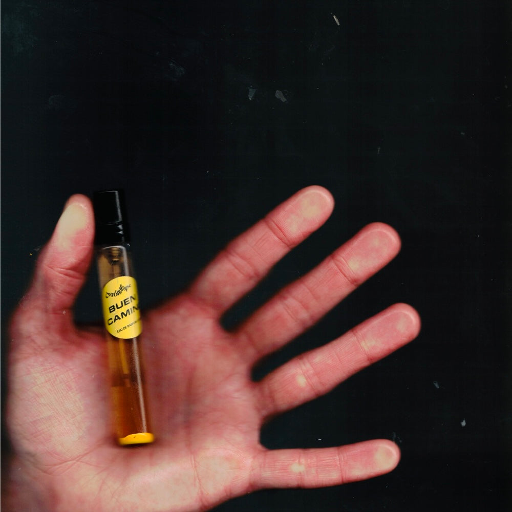 Hand holding a 7.5 mL travel spray of Chronotope Perfume’s Buen Camino Eau de Parfum, an avant-garde unisex fragrance of lavender, benzoin, and incense with herbal, resinous warmth, bandage and asphalt accords, soft almond sweetness, and lightly spiced citrus.