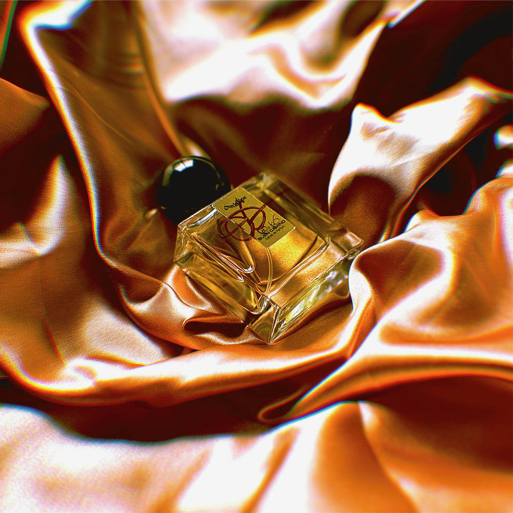 A bottle of Buen Camino Extrait de Parfum by Chronotope Perfume, nestled into shimmering gold fabric that reflects the color of the gold label on the face of the bottle.