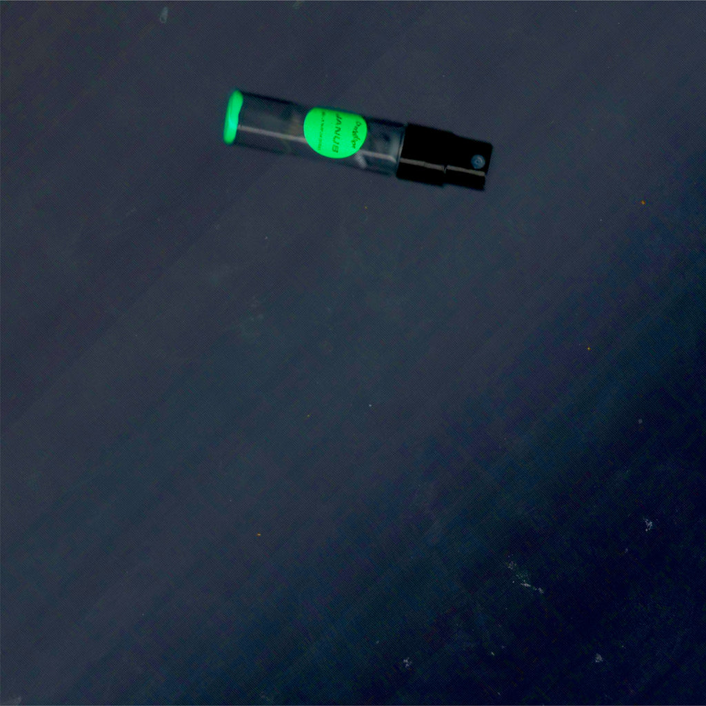 2mL spray sample of Chronotope Perfume’s Janus Eau de Parfum with fluorescent green label and matching enameled tip, resting on a steel-gray background.