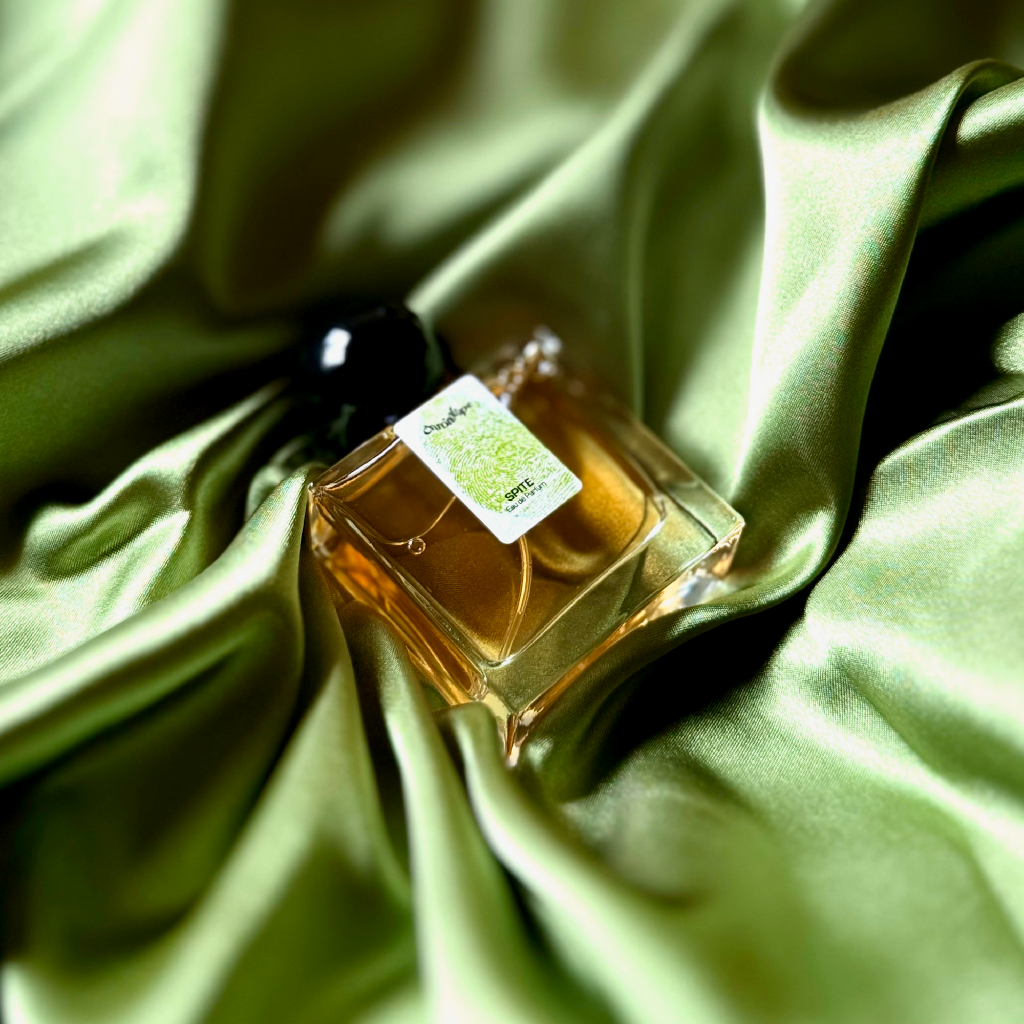 Nestled in a nest of green, silky, shining fabric with brilliant highlights and folds that appear black is a 30mL bottle of Chronotope Perfume’s Spite Eau de Parfum: A Perfume about Adaptation.