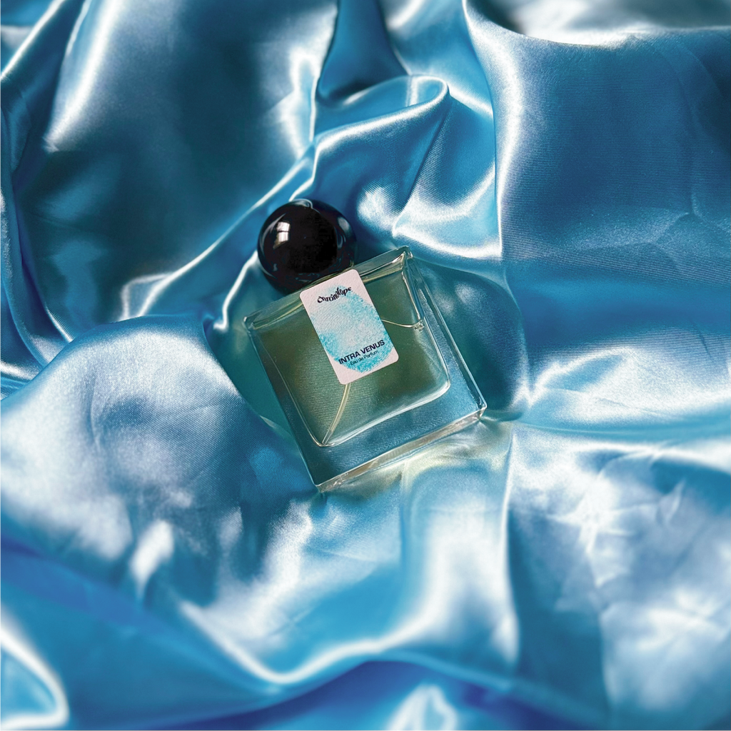 A full, 30mL bottle of Intra Venus Eau de Parfum by Chronotopeis nestled into a nest of colored, silky, shining fabric that matches the color of the thumbprint on the bottle’s label.
