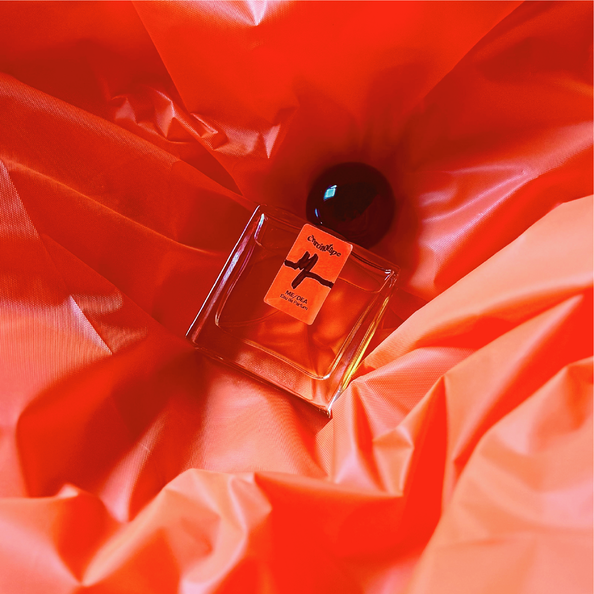 A 30mL bottle of Me/dea Eau de Parfum by Chronotope Perfume is nestled into a background of crinkled, fluorescent orange vinyl in an image showcasing the physical product.