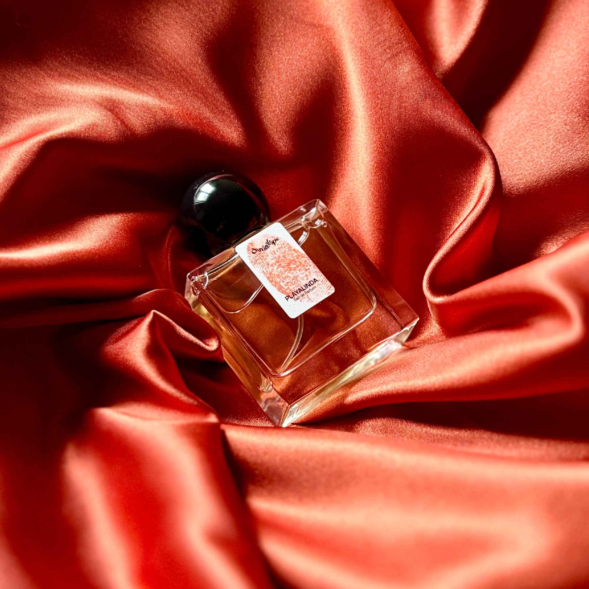 A 30mL bottle of Playalinda Eau de Parfum by Chronotope Perfume. It is nestled in shiny, silky fabric that matches the burnt orange color of the thumbprint on the bottle’s label. 